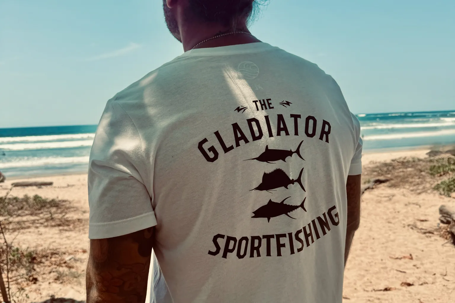 Persona con maglietta Gladiator Sportfishing sulla spiaggia, vista di spalle